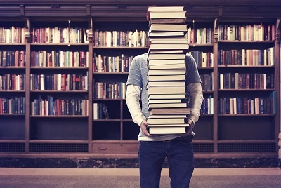 student with tall stack of books in library